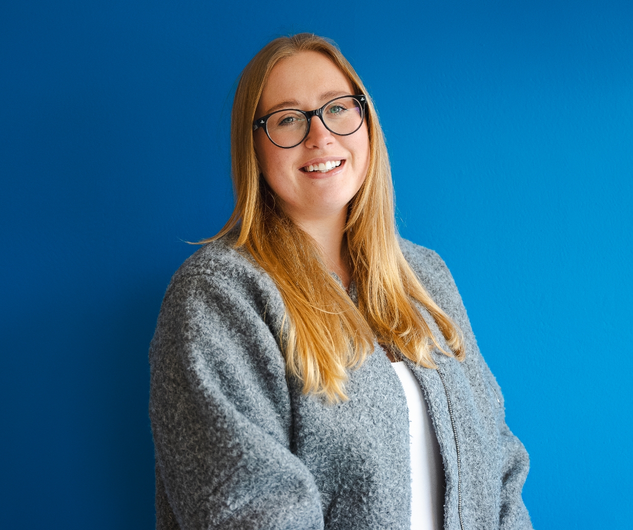 Tiff, an Account Manager, stands in a sweater and glasses before a blue wall.