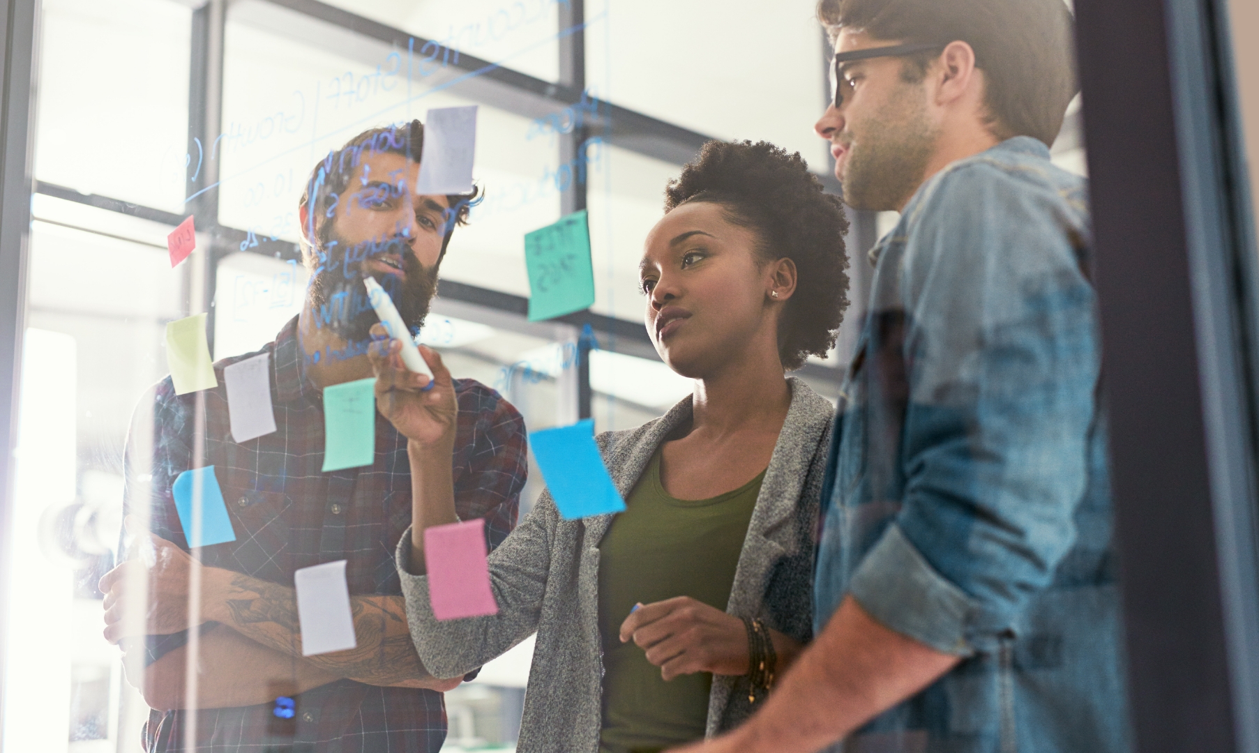Three people brainstorming in front of a glass wall covered with colorful sticky notes for strategy planning.