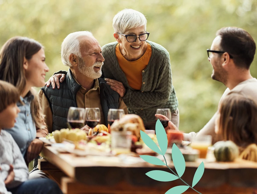 Three generations of a family gathered around an outdoor table enjoying food and drinks, smiling and talking in a nature setting.