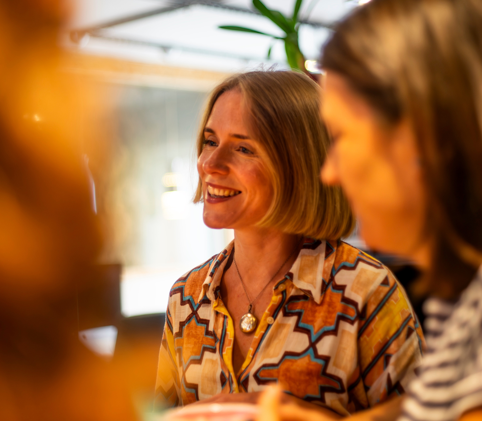 A woman with a blonde bob haircut smiles brightly while engaged in conversation, wearing a patterned shirt and a locket necklace, Halo Design Associates.