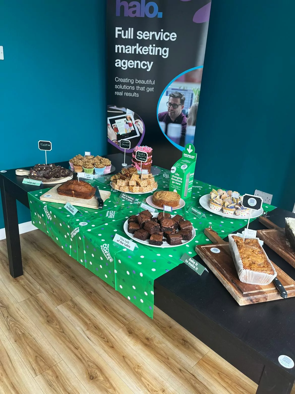 A bake sale table set up in front of a Halo marketing agency banner. The table is covered with a green polka dot tablecloth featuring the 'Macmillan Coffee Morning' logo. Various homemade baked goods, including brownies, cakes, and slices, are neatly arranged on plates and wooden boards. Small signs label the different treats. The Halo banner in the background promotes the agency as a 'Full service marketing agency,' with images of creative professionals at work.