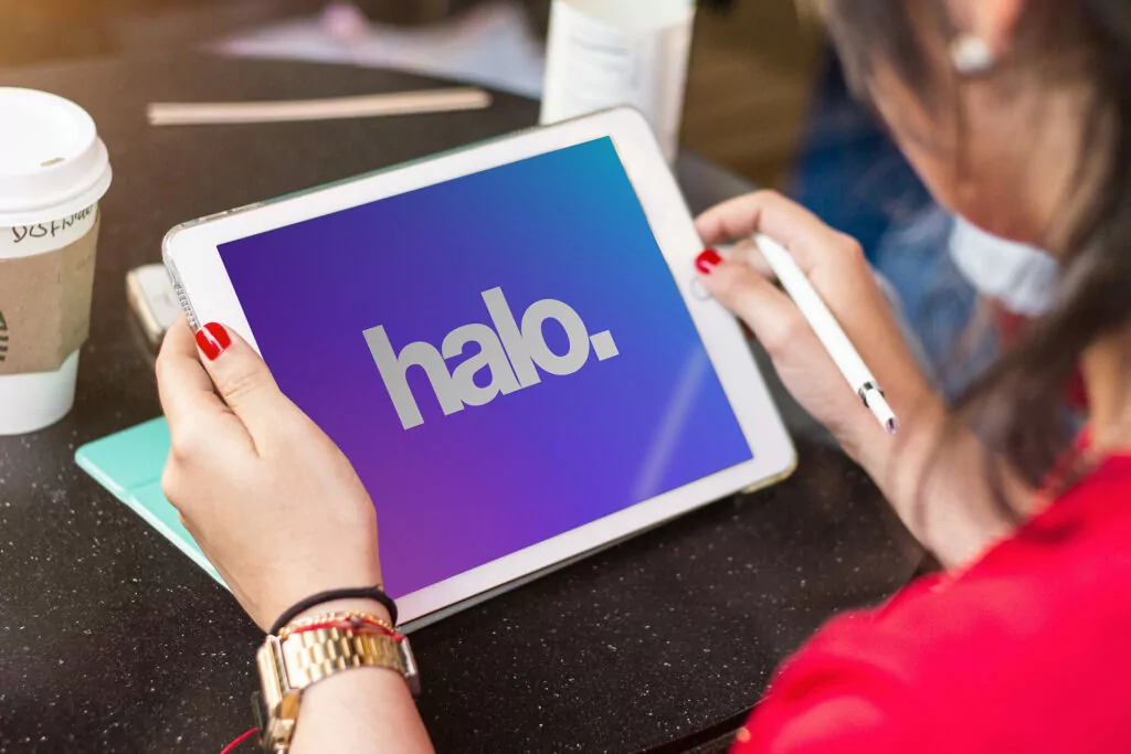 A woman holding a tablet with a stylus, displaying the word 'halo.' on a gradient blue and purple background. She is seated at a café table with a coffee cup nearby.