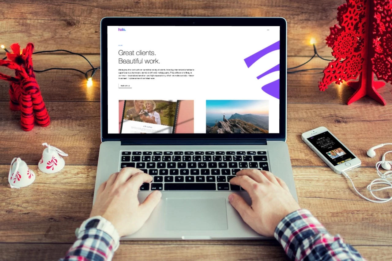 A person in a plaid shirt is typing on a laptop displaying a webpage with the 'halo.' branding. The webpage features a headline that reads 'Great clients. Beautiful work.' alongside images of a couple and a scenic mountain view. The workspace has a cozy wooden aesthetic with festive decorations, including a red wooden tree and hanging fairy lights. A smartphone with earphones plugged in rests on the table, displaying a streaming app.