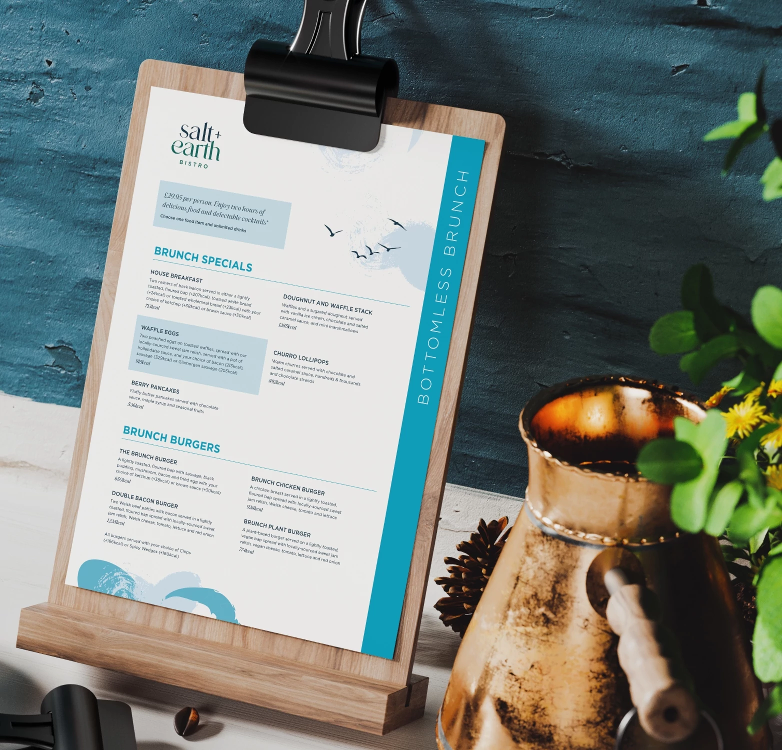 A stylishly designed bottomless brunch menu from Salt + Earth Bistro, displayed on a wooden clipboard against a rustic blue backdrop. The menu features a selection of brunch specials such as house breakfast, waffle eggs, berry pancakes, and churro lollipops, along with gourmet brunch burgers. A vintage copper pitcher and fresh greenery add to the cozy and inviting atmosphere of the setting.