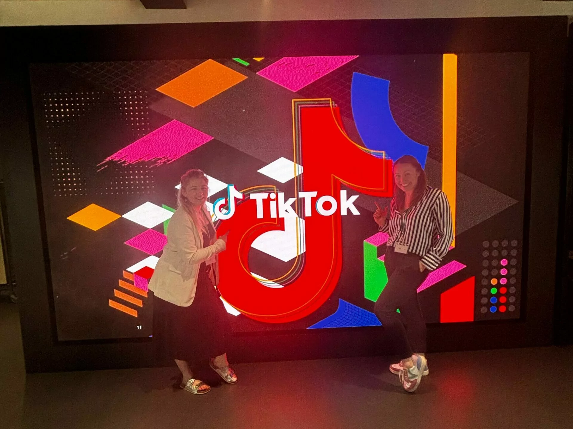 Two women posing and smiling in front of a large illuminated TikTok logo display with a colorful geometric background at what appears to be a TikTok event or venue.