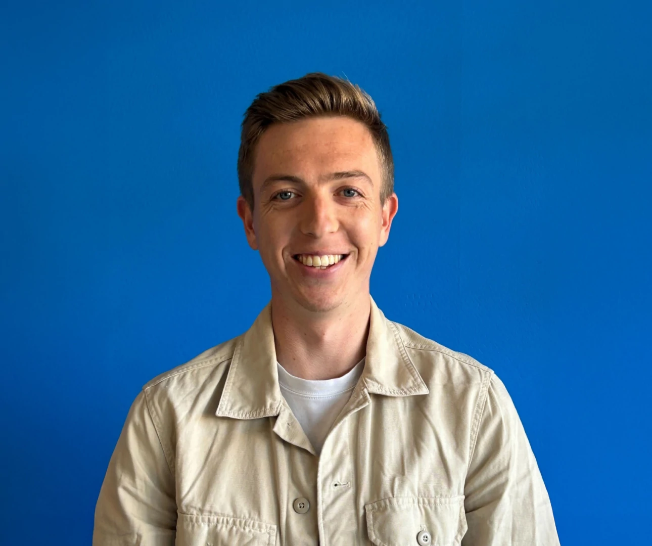 Oscar, an Account Manager, smiles at the camera while wearing a tan jacket against a blue wall backdrop.